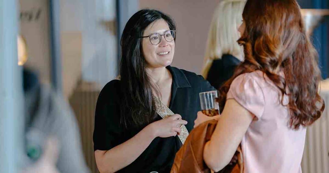 Two women stand facing each other and talk. One woman with glasses is smiling, holding a drink. The other woman, with long red hair, is seen from behind, holding a coat. The background is out of focus.
