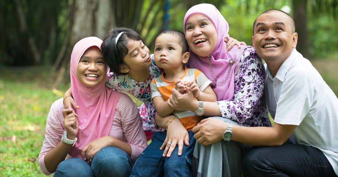 A joyful family sits together on grass in a park. Two women in pink headscarves, a girl kissing a toddler’s cheek, and a smiling man all pose closely, exhibiting a sense of togetherness and happiness amidst lush greenery.