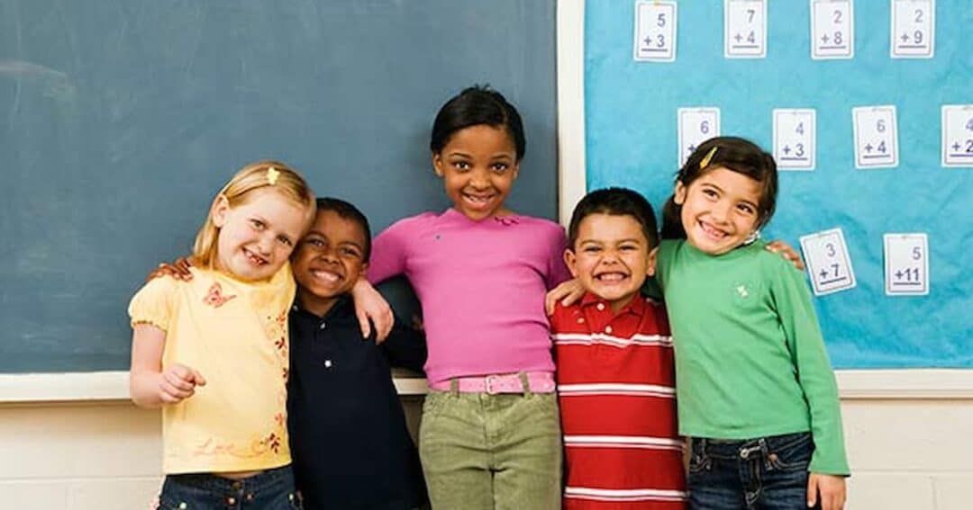 Five children pose together in front of a chalkboard and a wall with math flashcards. They are smiling and have their arms around each other, standing in a classroom setting.
