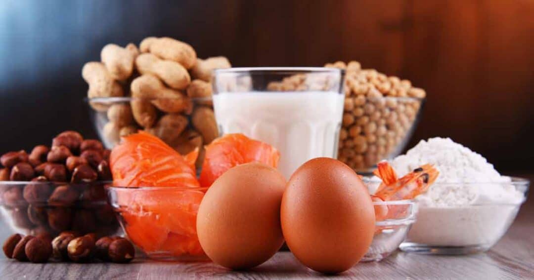 Various foods are displayed, including peanuts, milk, soybeans, hazelnuts, salmon, shrimp, and eggs, arranged on a wooden surface. These items are commonly associated with food allergies.