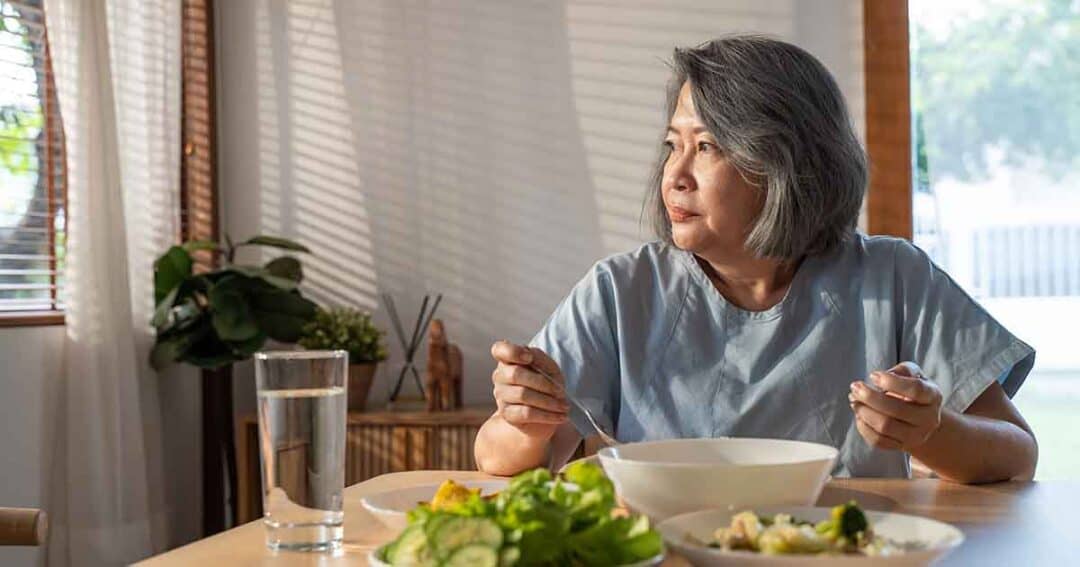 A woman with gray hair sits at a dining table, looking thoughtfully out the window. There are plates of salad and a bowl in front of her. A glass of water is on the table, and sunlight filters through the window blinds.