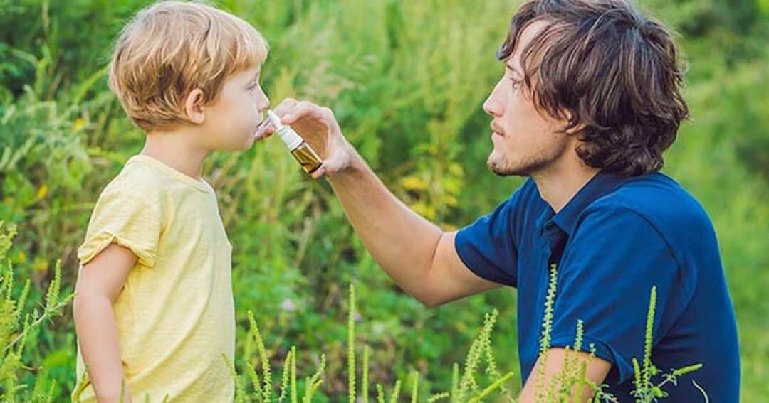 A man kneels on the grass while giving a young child a drink from a small cup. The child is wearing a yellow shirt, and the man is in a blue shirt. They are surrounded by greenery, with plants and grass in the background.