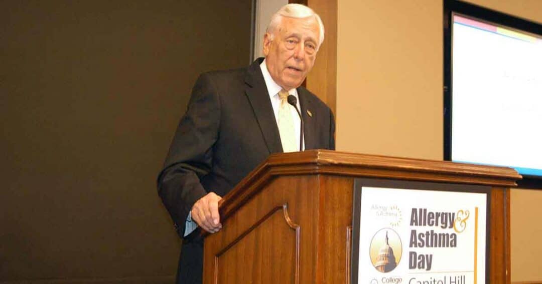 Steny Hoyer speaks at a podium labeled “Allergy & Asthma Day on Capitol Hill.” A projection screen and brown walls are in the background.