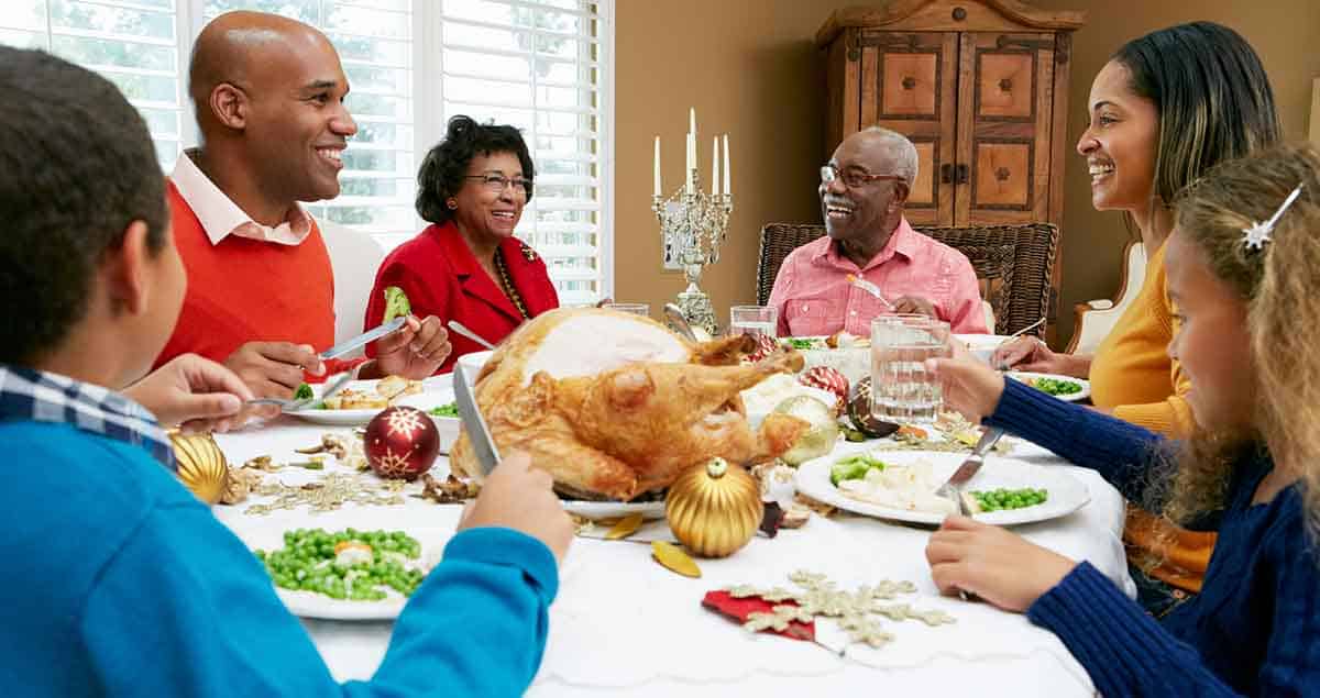A family gathered around a dining table enjoying a holiday meal. The table is set with a roast turkey, decorative ornaments, and side dishes. Everyone is smiling and engaging in conversation.